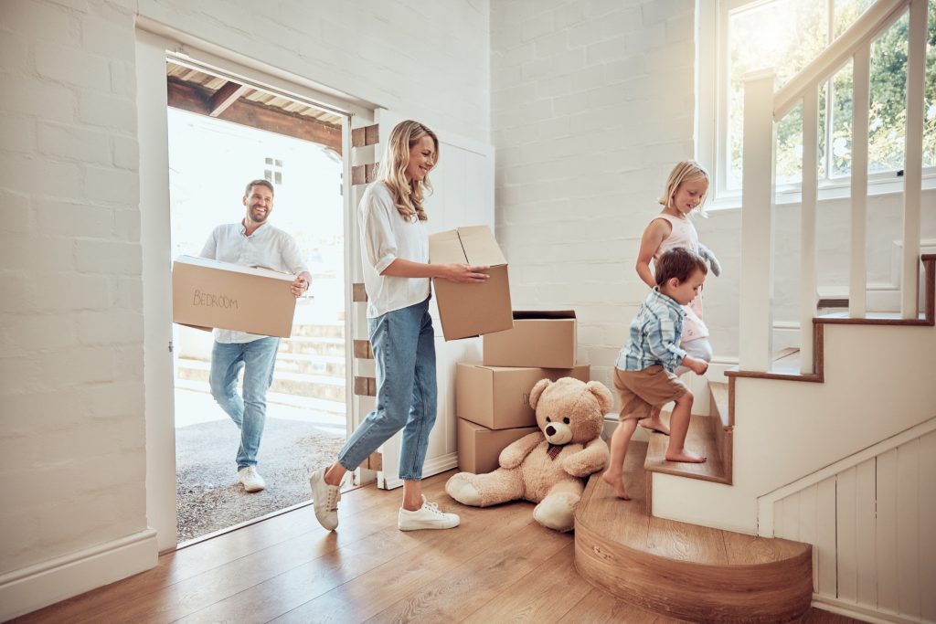 happy caucasian parents with little kids moving into their new home. excited couple carrying boxes up staircase with their son and daughter. adorable children helping to unpack and move into property